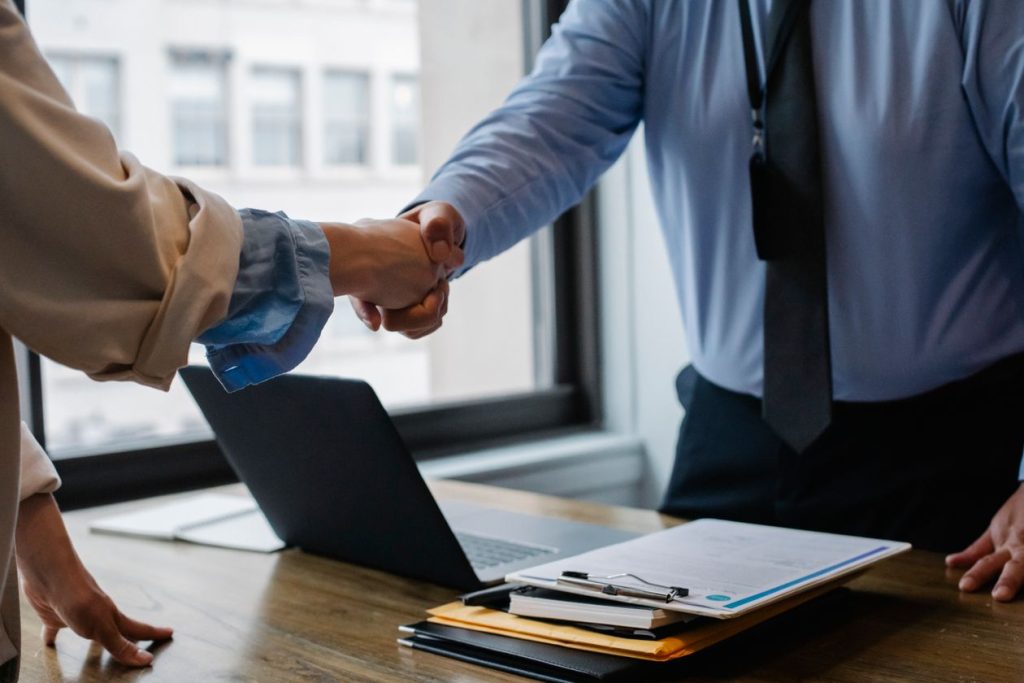 Business owner shaking hands with a returning client across a desk with a laptop and clipboard, illustrating new versus existing customer classification