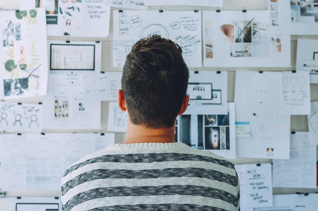 Man viewed from behind studying a wall of printed storyboards and layout mockups, illustrating the creative planning a fifteen-second video ad demands