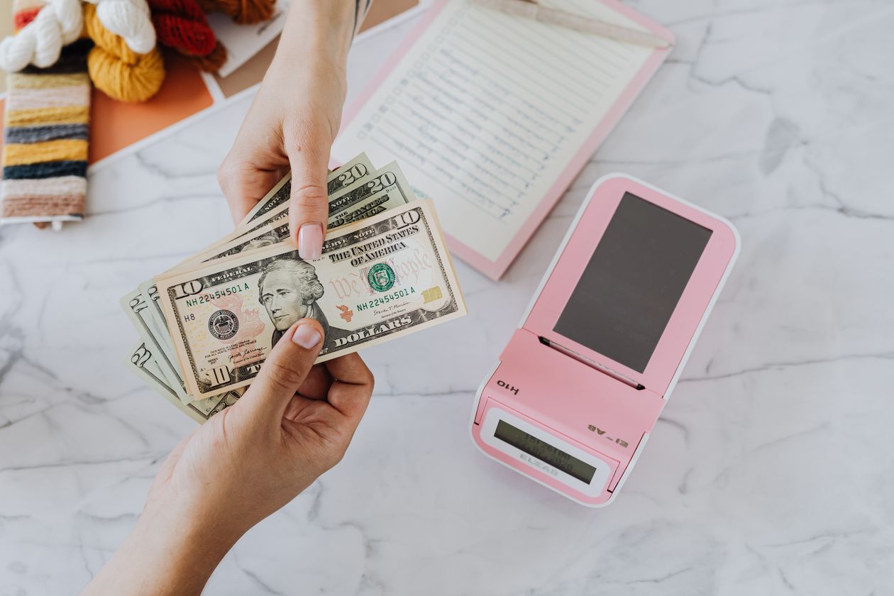 Hands counting cash beside a card terminal on a marble counter, illustrating a small business weighing a new monthly platform subscription cost