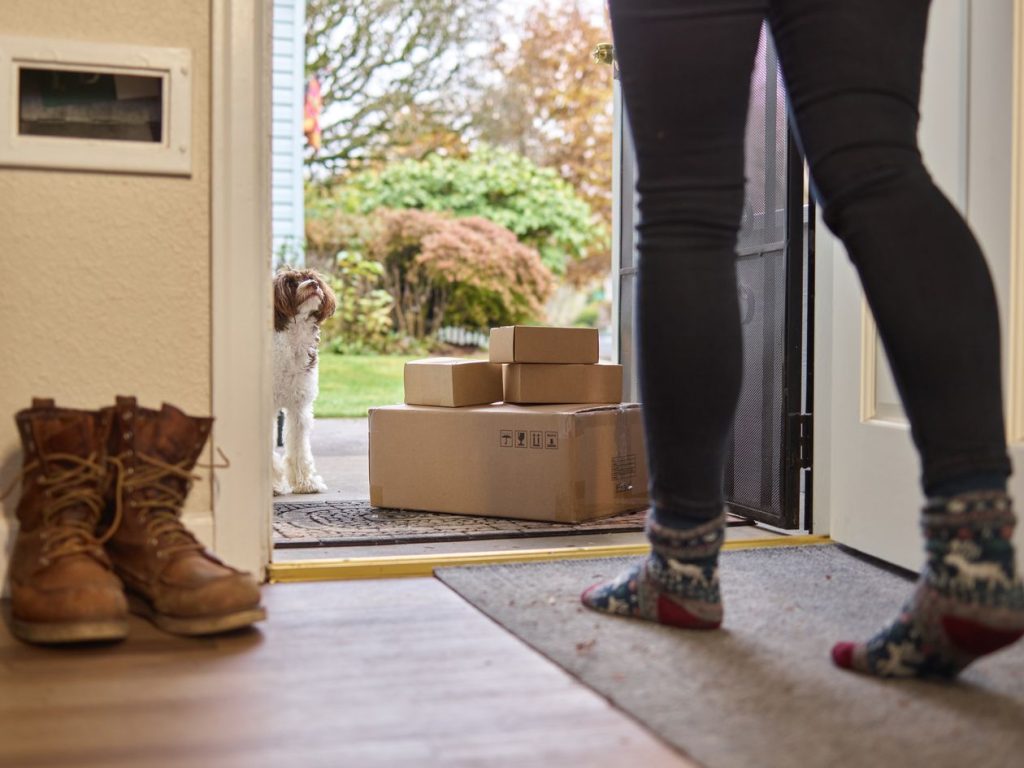 Customer in wool socks opening her front door to stacked cardboard delivery boxes and a curious dog, the post-purchase moment that produces store reviews