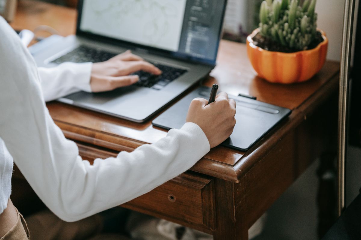 A designer works with a stylus, graphics tablet and laptop at a wooden desk.