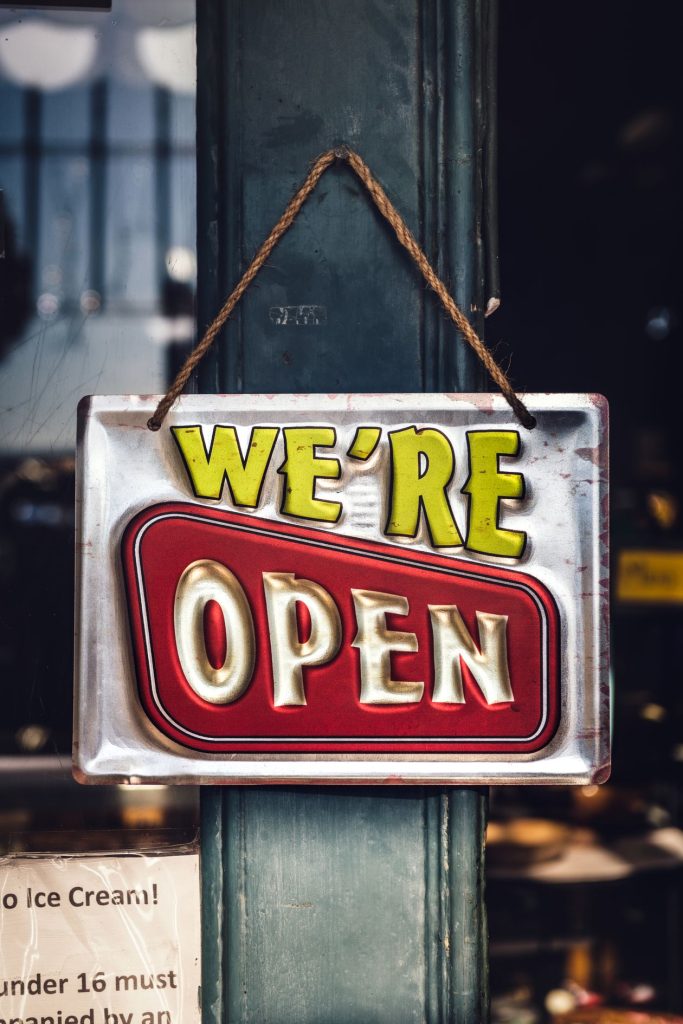 Metal "We're Open" sign hanging by a rope on a shop doorway pillar, illustrating the business name local customers recognize before they click an ad
