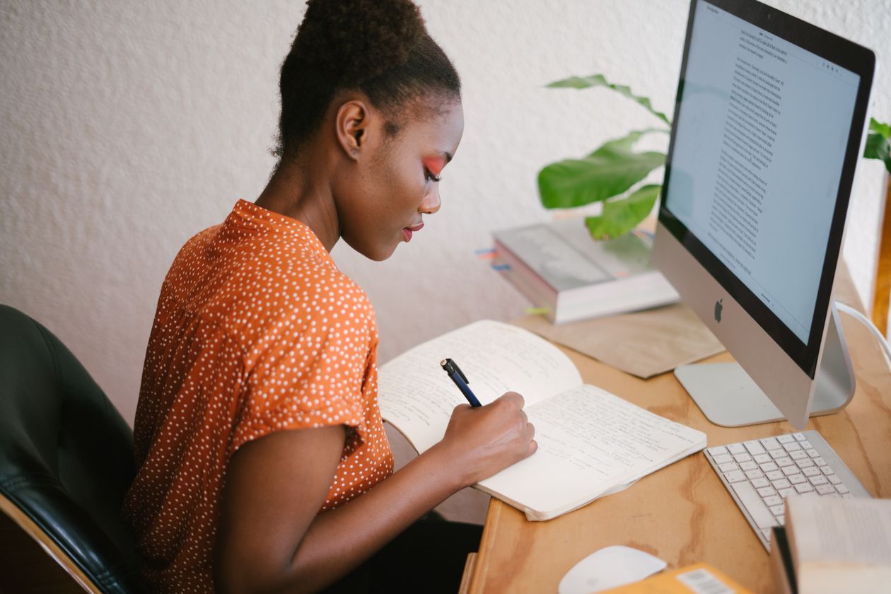 Woman writing notes in a notebook beside a desktop monitor showing a text document, illustrating a real author drafting a small business blog post