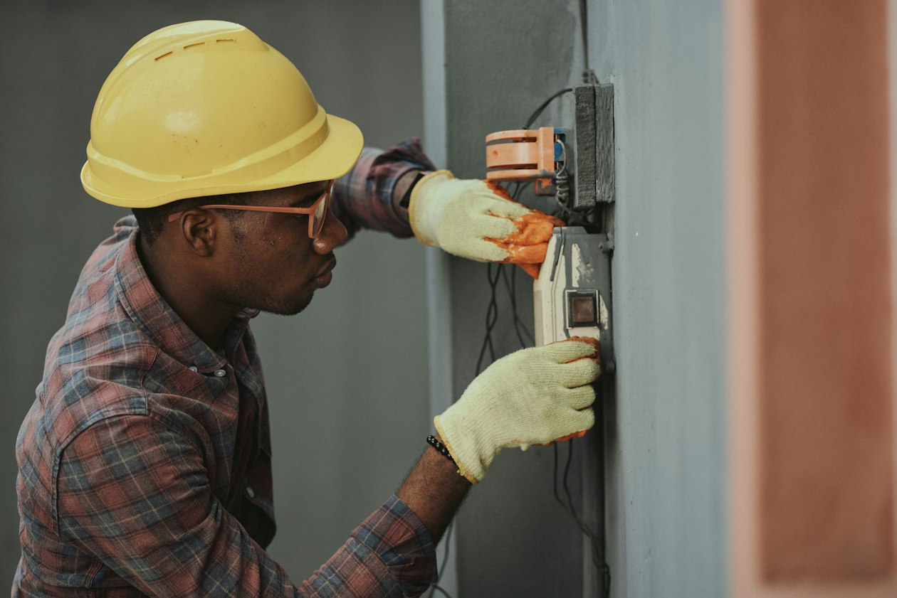 Electrician in a yellow hard hat testing a wall electrical panel with a meter, illustrating a local service business qualifying leads before sending a technician